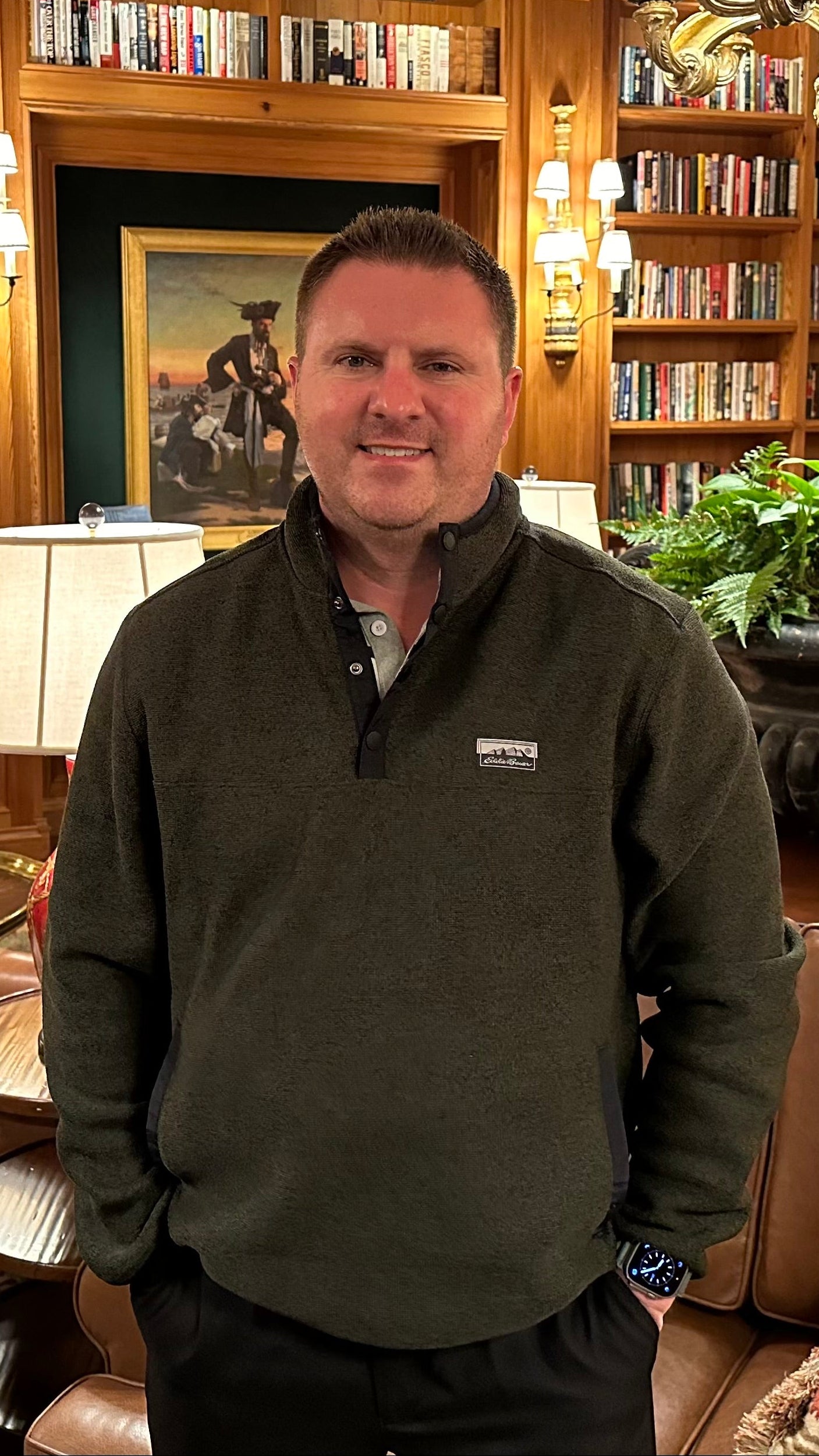 Man standing in a library with bookshelves, a chandelier, and a painting on the wall.
