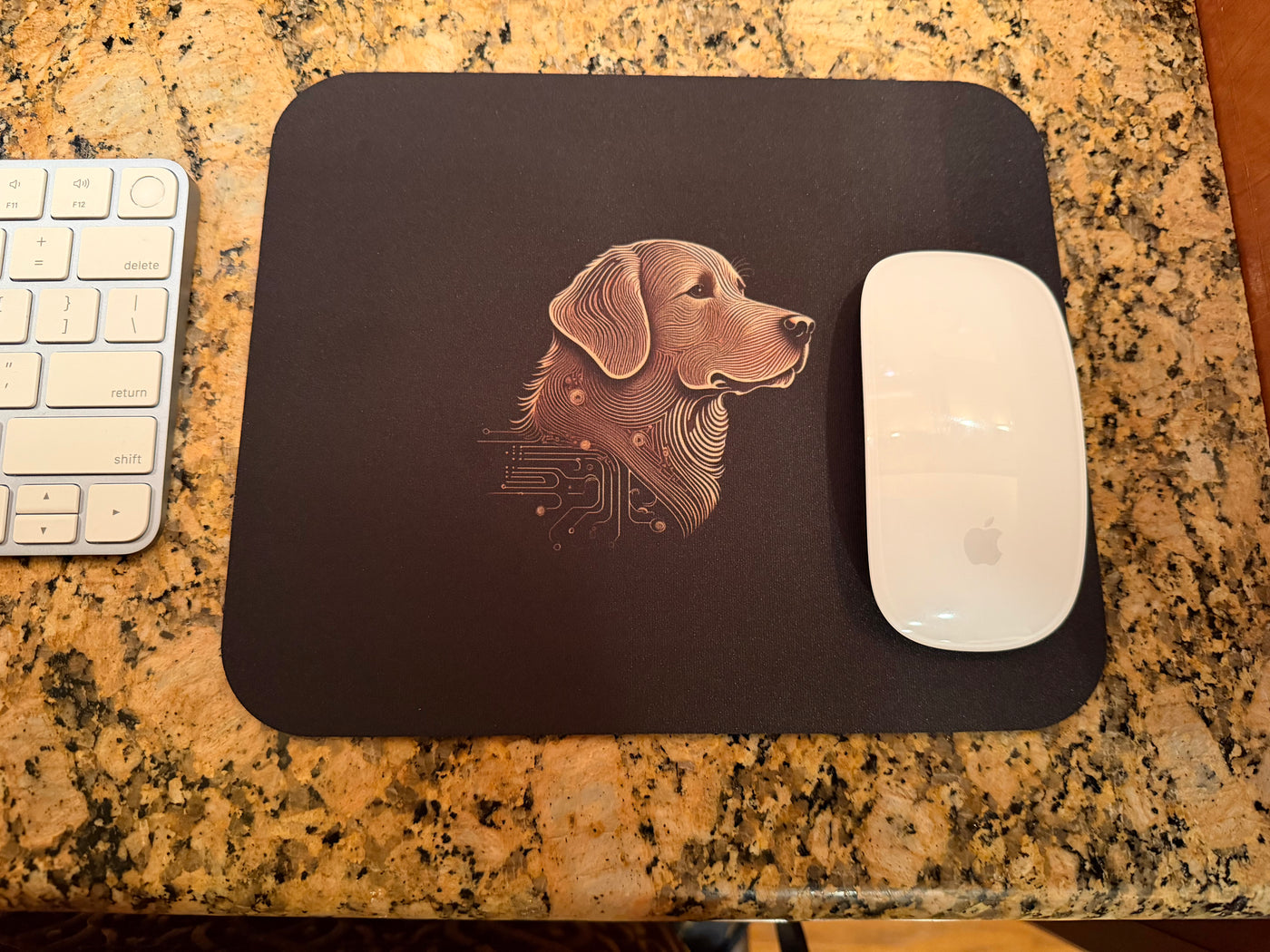Black rectangular mouse pad with stylized dog profile print beside white Apple mouse and keyboard