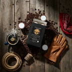 Coffee beans, coffee cup, candles, leather gloves, and rope on a wooden surface with a Byrd & Dark Brew coffee package.